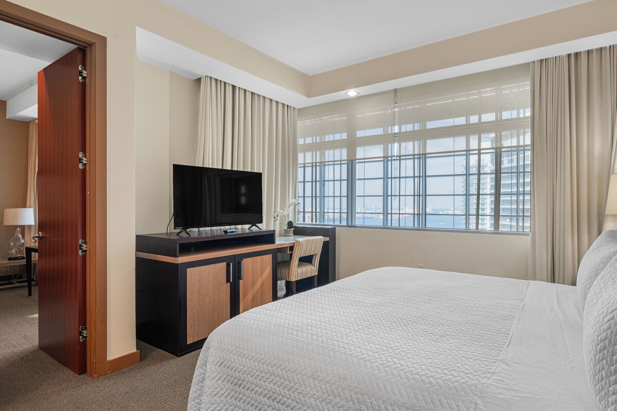 Habitación de hotel con cama, escritorio con televisor, ventana con cortinas y vista a un paisaje urbano.