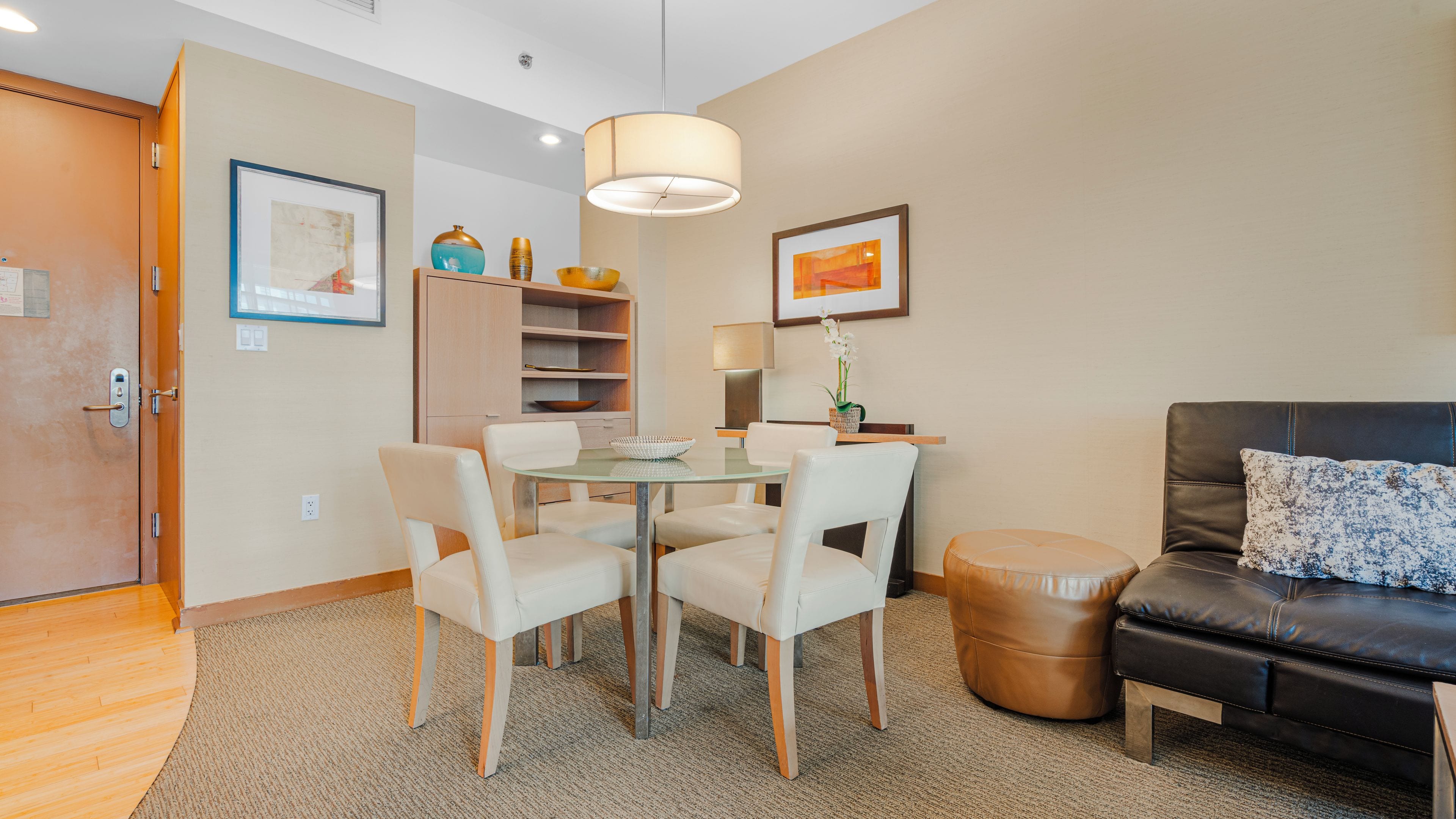 Un salon moderne avec une table à manger en verre, des chaises blanches, un canapé en cuir noir et un pouf marron.