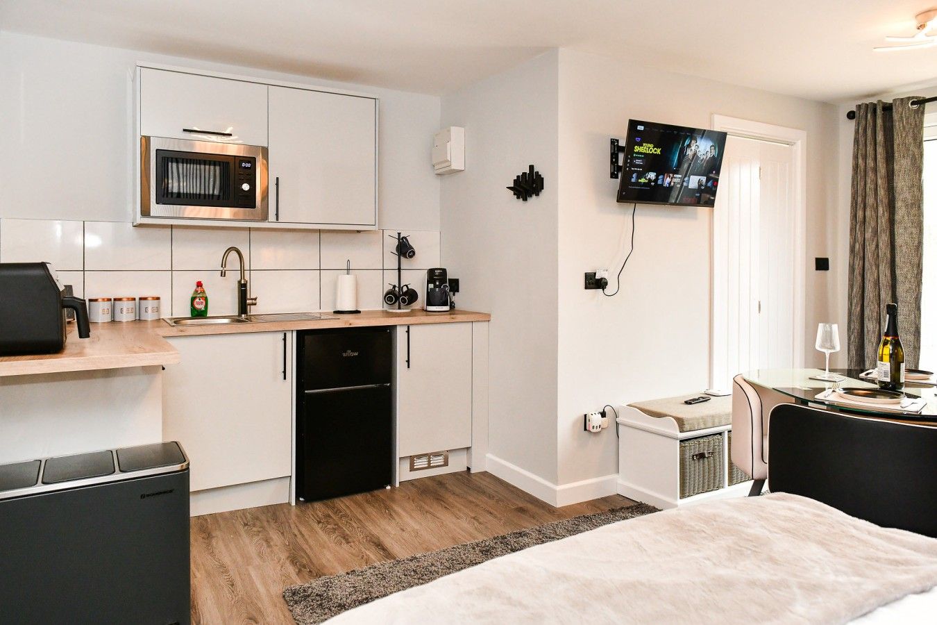 A compact hotel room with a kitchenette, a bed, a TV mounted on the wall, and a small seating area.