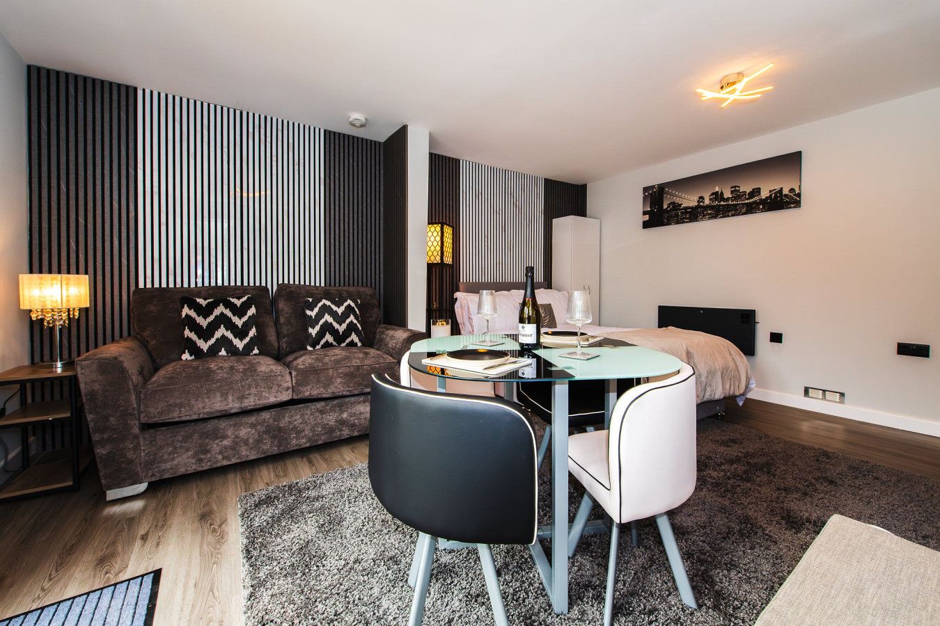 A modern living room with a sofa, a dining table with chairs, a lamp, and a cityscape painting on the wall.