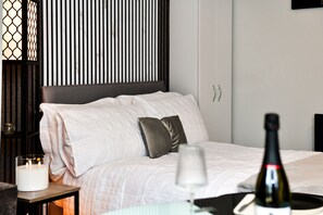 Hotellihuone, jossa on sänky, yöpöytä kynttilällä, pullo viiniä ja seinälle asennettu televisio.