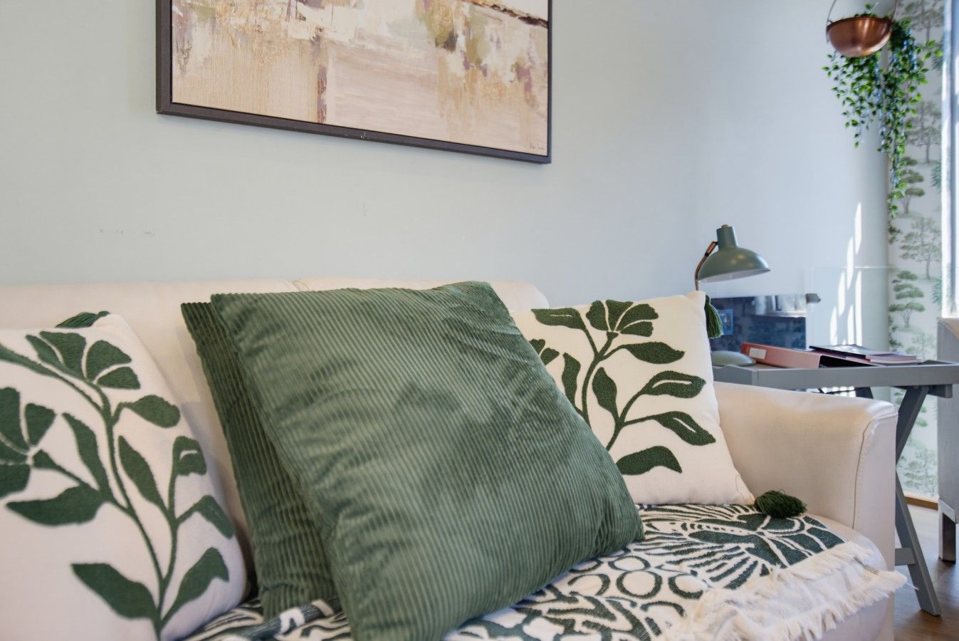 Een zithoek met een bank, een groen kussen en een gebogen plaid. Aan de muur hangt een schilderij en van het plafond hangt een plant.