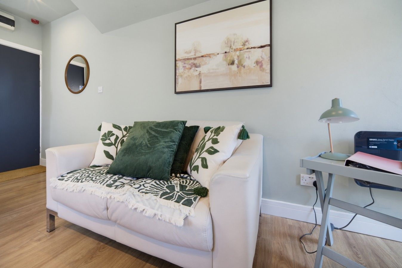 A modern living room with a white sofa, green and white patterned throw, a round mirror, a painting, and a desk with a lamp and a printer.