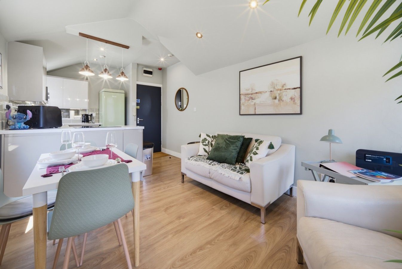 A modern living room with a white sofa, a dining table set for two, and a kitchen in the background.