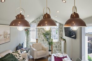 Ein modernes Wohnzimmer mit hohen Decken, drei Pendelleuchten, einem Sofa, einem Sessel und einem Esstisch für zwei Personen.