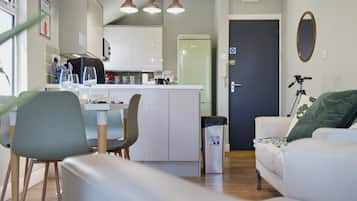 Ein modernes Wohnzimmer mit Sofa, Esstisch und Stühlen. Im Hintergrund ist eine Küchenzeile mit Kühlschrank und Schränken zu sehen.
