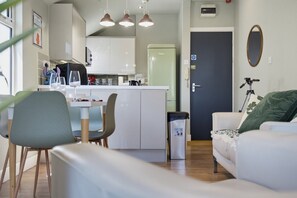 Ein modernes Wohnzimmer mit Sofa, Esstisch und Stühlen. Im Hintergrund ist eine Küchenzeile mit Kühlschrank und Schränken zu sehen.