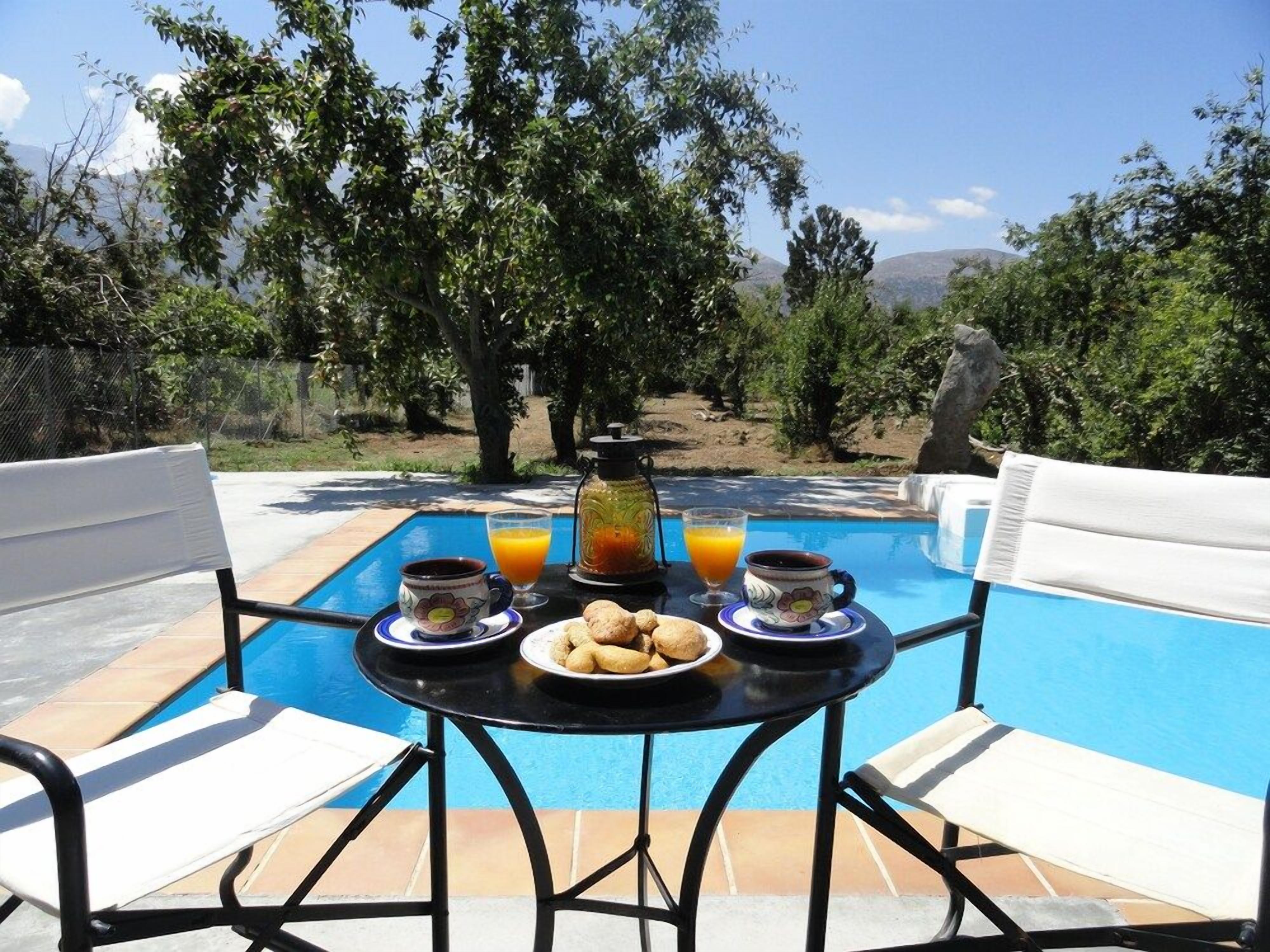 Mesa junto a la piscina con alimentos para el desayuno, dos sillas y vistas a árboles y montañas.