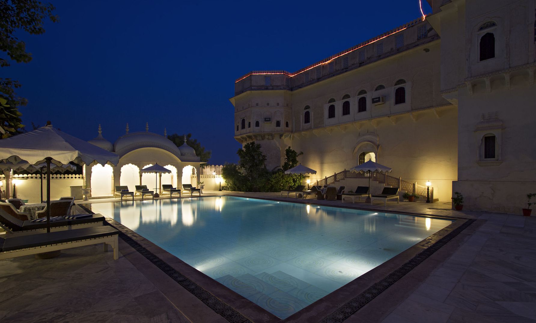 A pool area with lounge chairs and umbrellas, surrounded by a building with arches and a dome.