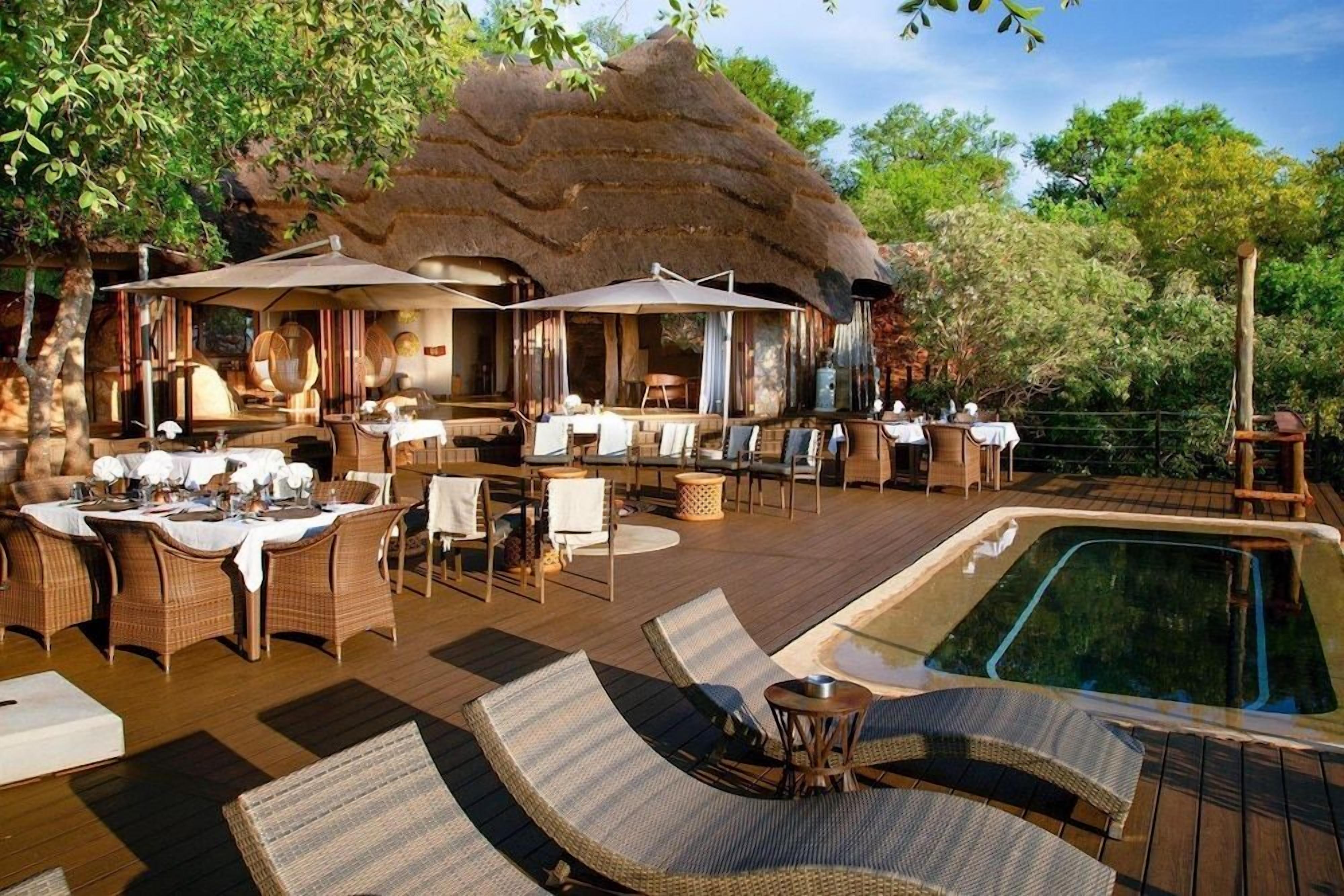 An outdoor dining area with tables and chairs, a pool, and a thatched-roof structure.