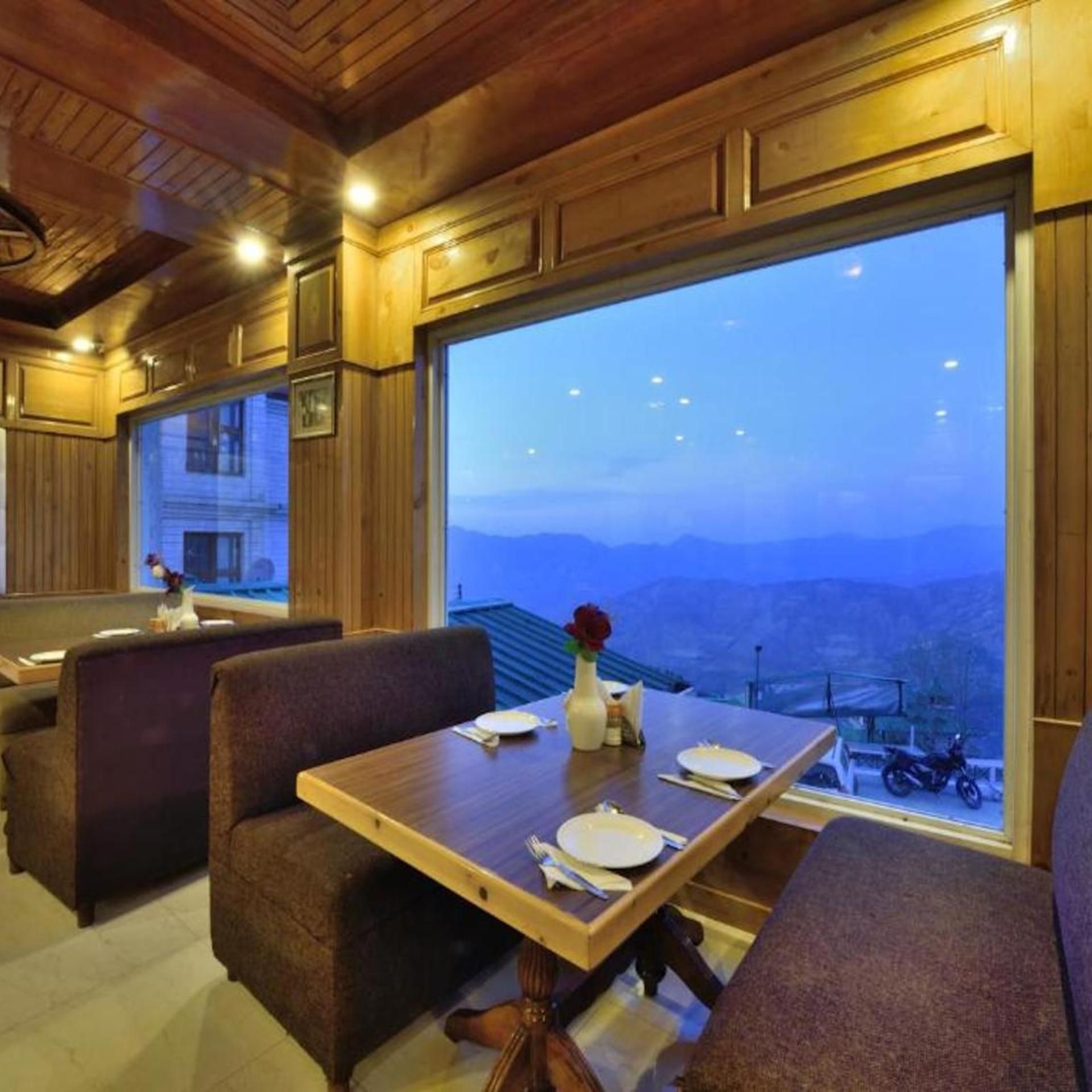 A dining area with a table set for two, a vase with a flower, and a view of mountains through large windows.