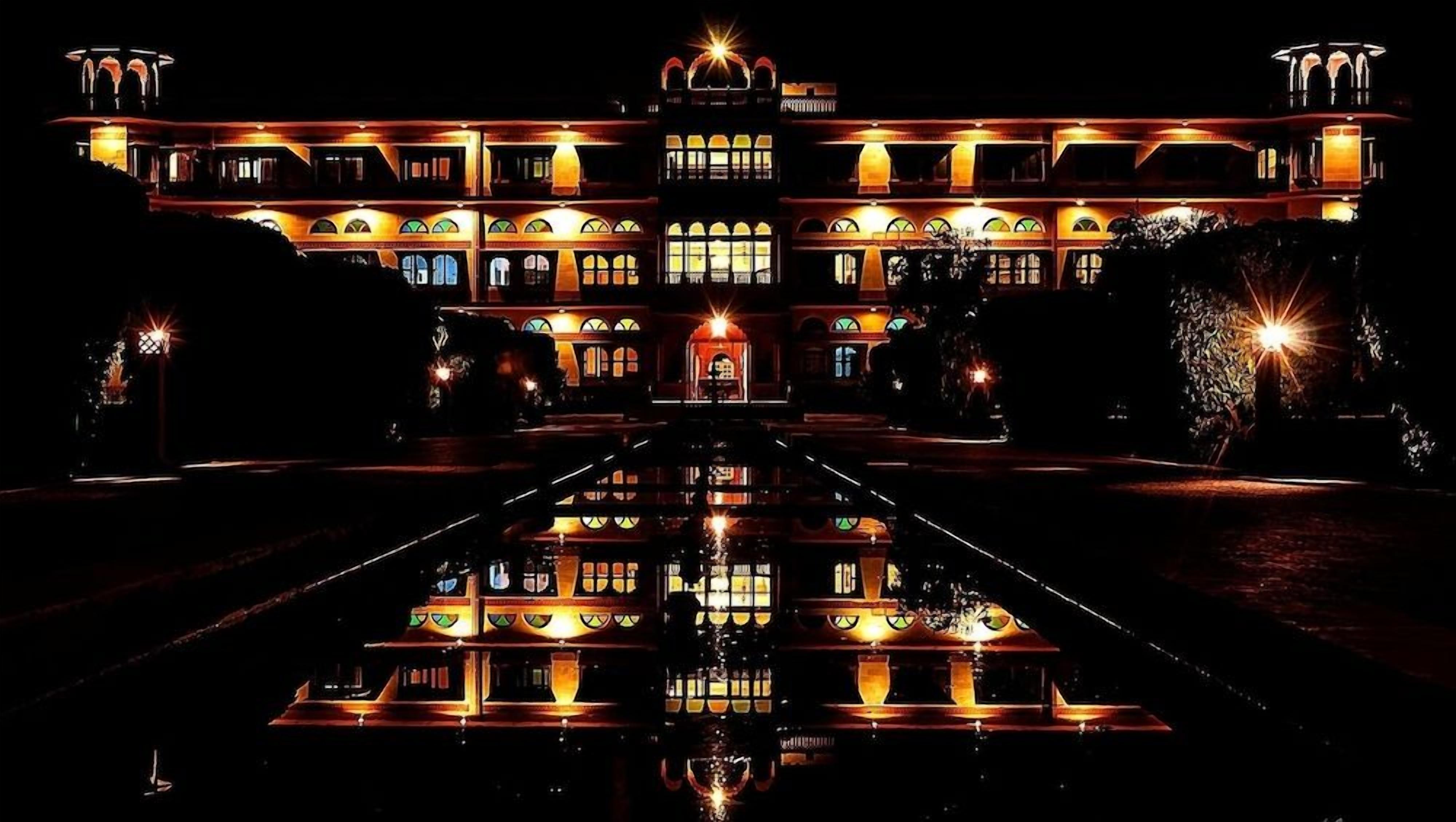 A large, illuminated building with a symmetrical facade, a central entrance, and a reflective water feature in front.