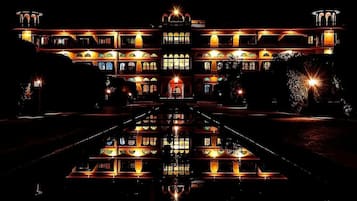 Ein großes, beleuchtetes Gebäude mit einer symmetrischen Fassade, einem zentralen Eingang und einem spiegelnden Wassenspiel davor.