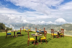 Outdoor dining tables with chairs on a grassy hill with a mountain view. - Kufri Holiday Resort (Theog)