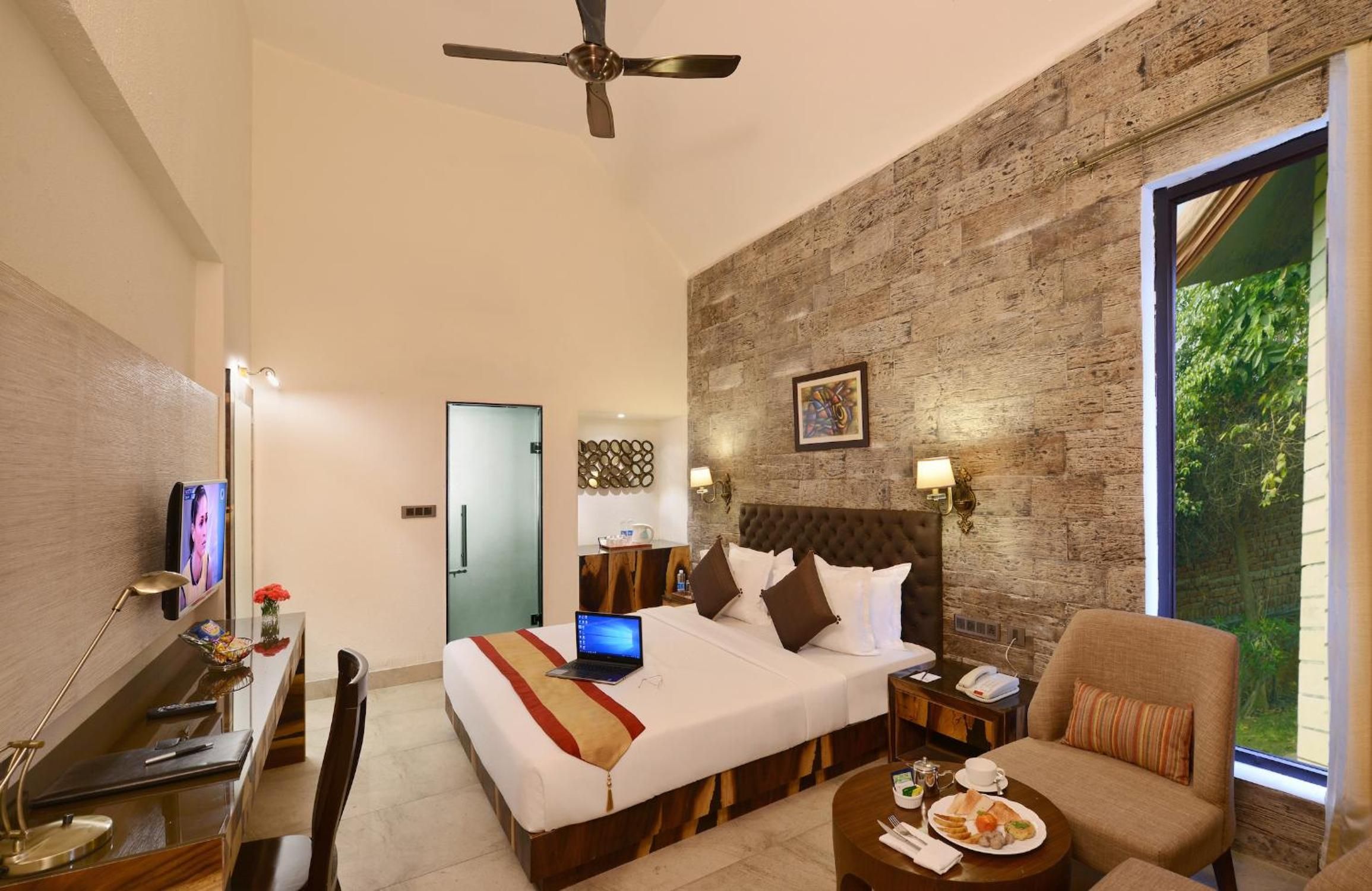 A hotel room with a bed, a desk with a laptop, a chair, a dining table with breakfast, and a large window.
