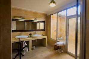 A bathroom with two sinks, a large mirror, a shower, and a view through the glass door. - Fort Khejarla (Bilara)