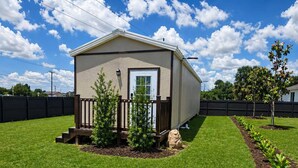 A single-story mobile home with a front porch, a small garden area with young trees, and a clear blue sky. - Modern Independent Tiny House (Lehigh Acres)