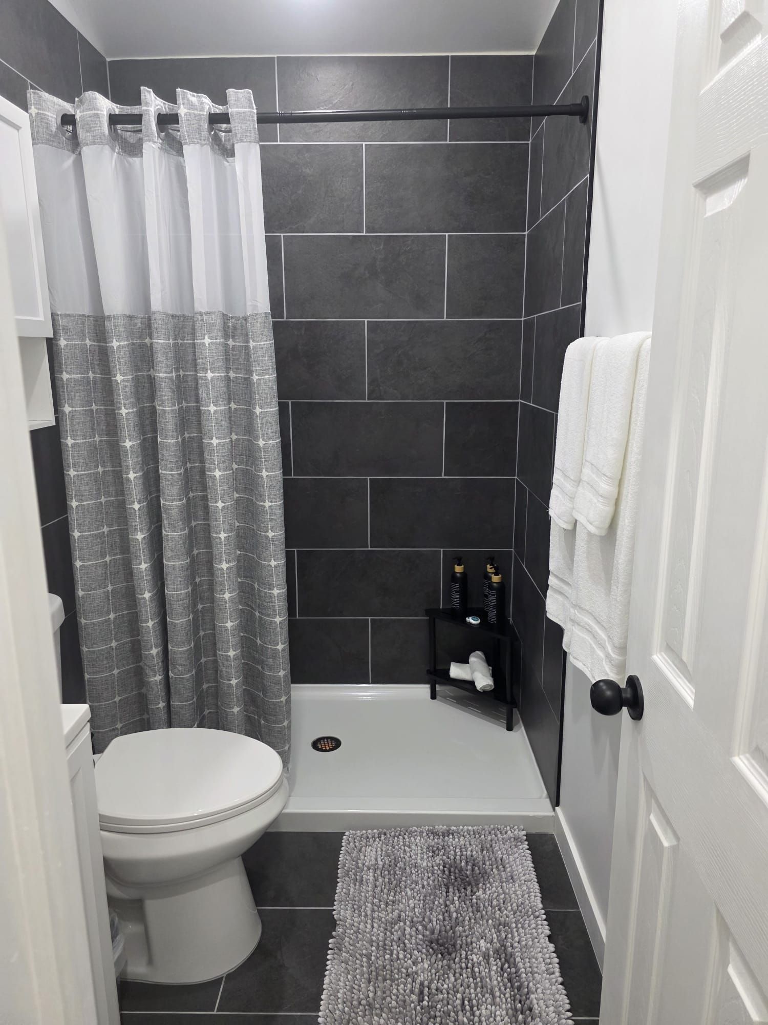 Ein Badezimmer mit einer weißen Toilette, einer Dusche mit grauem, gemustertem Vorhang, einem schwarzen Ablagefach für Toilettenartikel und einem weißen Handtuchhalter.
