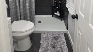 A bathroom with a white toilet, a shower with a grey patterned curtain, a black shelf with toiletries, and a white towel rack. - Modern Independent Tiny House (Lehigh Acres)