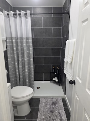 Un baño con un inodoro blanco, una ducha con cortina gris estampada, un estante negro con artículos de tocador y un perchero blanco.