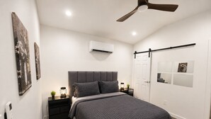 A modern bedroom with a bed, nightstands, a ceiling fan, an air conditioning unit, and artwork on the walls. - Modern Independent Tiny House (Lehigh Acres)
