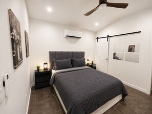Un dormitorio moderno con cama, mesitas de noche, ventilador de techo, equipo de aire acondicionado y obras de arte en las paredes.
