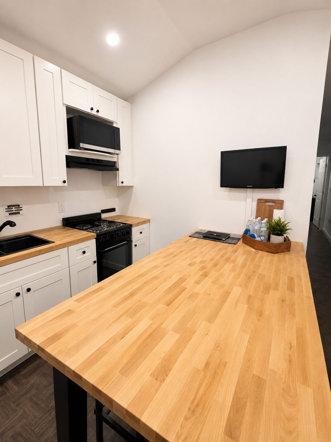 A modern kitchen with white cabinets, a wooden countertop, a stove, and a flat-screen TV mounted on the wall.
