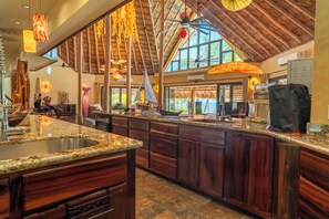 A spacious kitchen with a large island, wooden cabinets, and a view of the ocean through large windows. - Casa Pisces-Deluxe Beachfront Mansion (San Pedro)