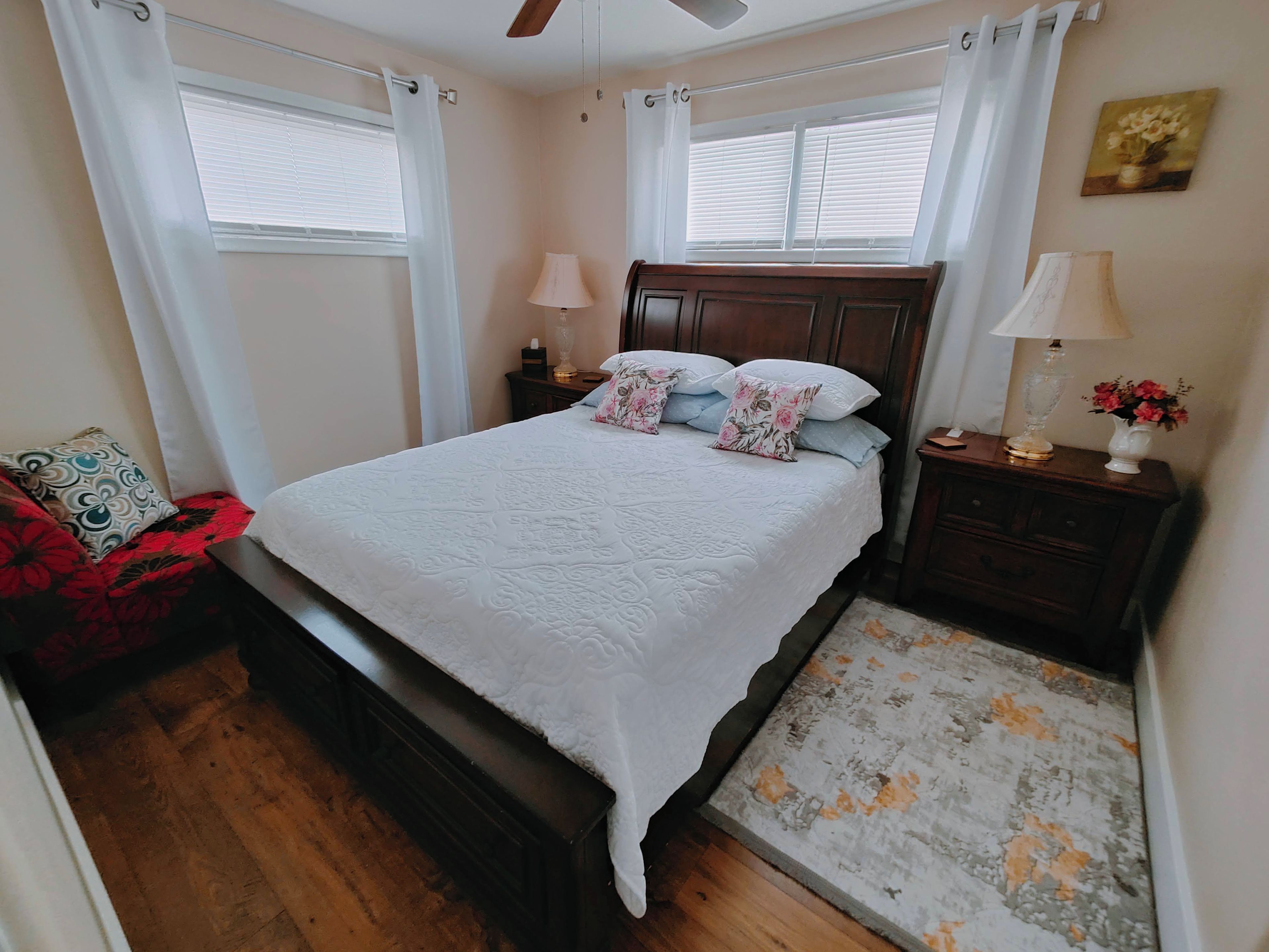 A bedroom with a bed, nightstands, a wooden headboard, a red chair, a wooden floor, and a window with curtains.