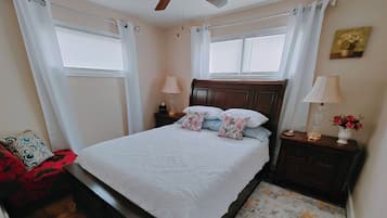 A bedroom with a bed, nightstands, a wooden headboard, a red chair, a wooden floor, and a window with curtains.