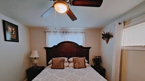 Een slaapkamer met een bed, nachtkastje, lamp, plafondventilator, raam met zonwering en een wandplant.