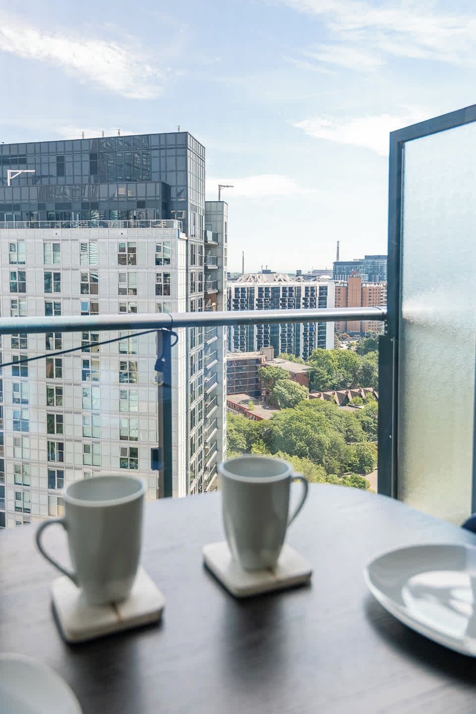 A balcony with two white cups on coasters, overlooking a cityscape with modern buildings and trees.