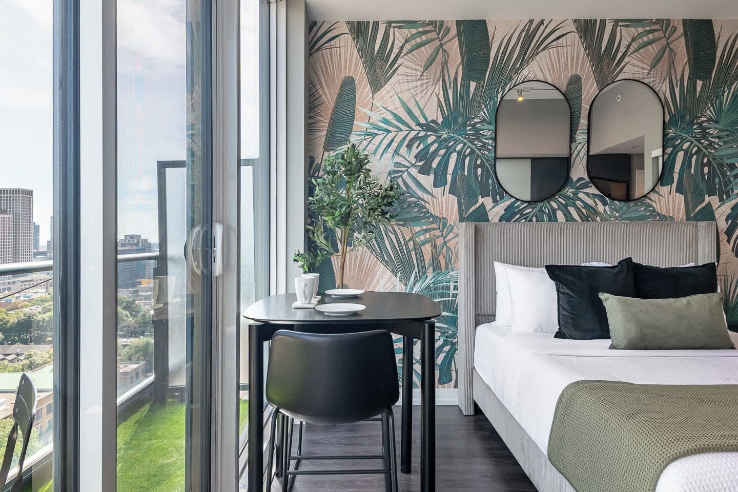 A hotel room with a large window, a bed with multiple pillows, a round table with a chair, and a tropical leaf wallpaper.