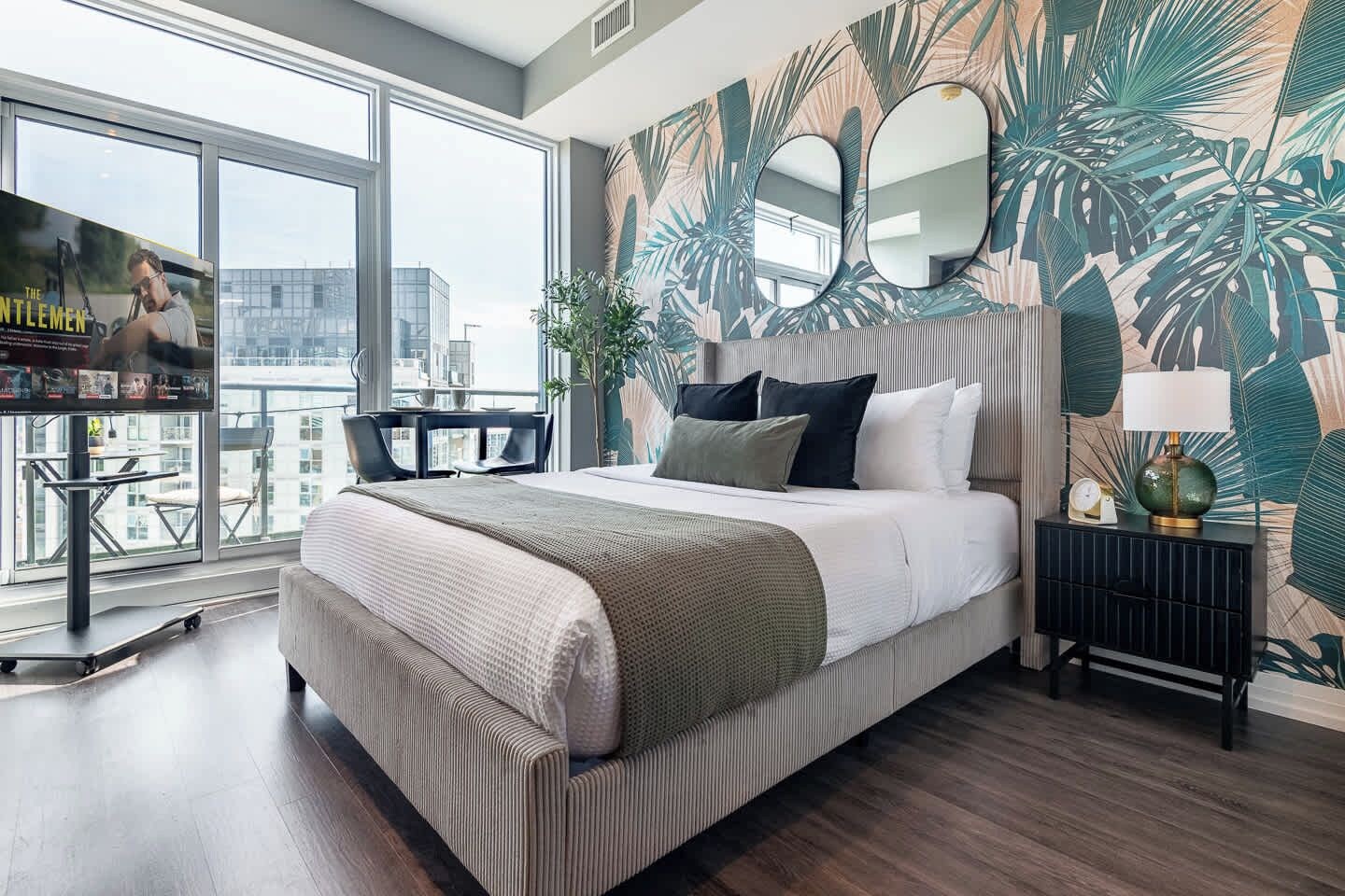 Una habitación de hotel moderna con una cama grande, un balcón con vistas, un televisor y un papel tapiz tropical.