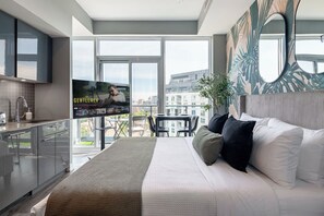 Ein modernes Hotelzimmer mit einem großen Bett, einem Fernseher, einem Balkon mit Blick auf die Stadt und einer Küchenzeile.