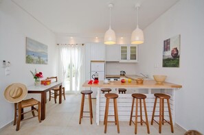 A modern kitchen with white cabinetry, a central island with stools, pendant lighting, and artwork on the walls. - Kavos Boutique Hotel Naxos (Naxos)