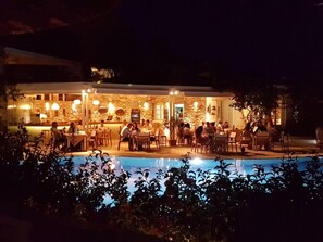 Un área de comedor exterior de noche con paredes de piedra y mesas, rodeada por una piscina.