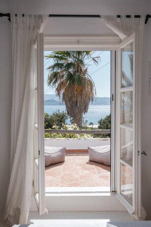 Una habitación con un ventanal grande, cortinas blancas y vistas a una palmera y al mar.