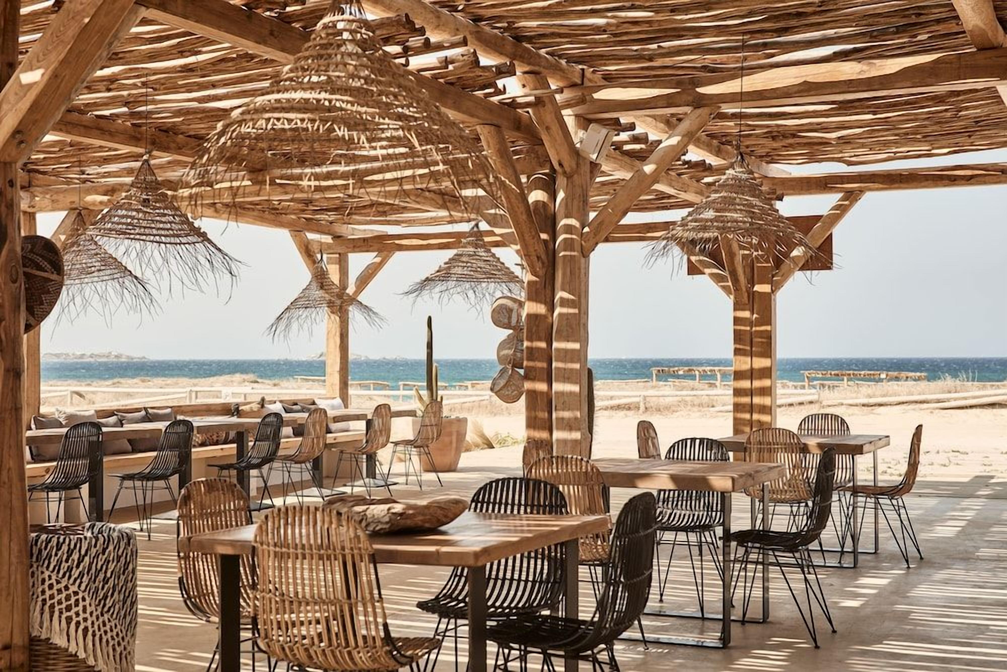 An outdoor dining area with wooden tables and chairs, thatched umbrellas, and a view of the beach.