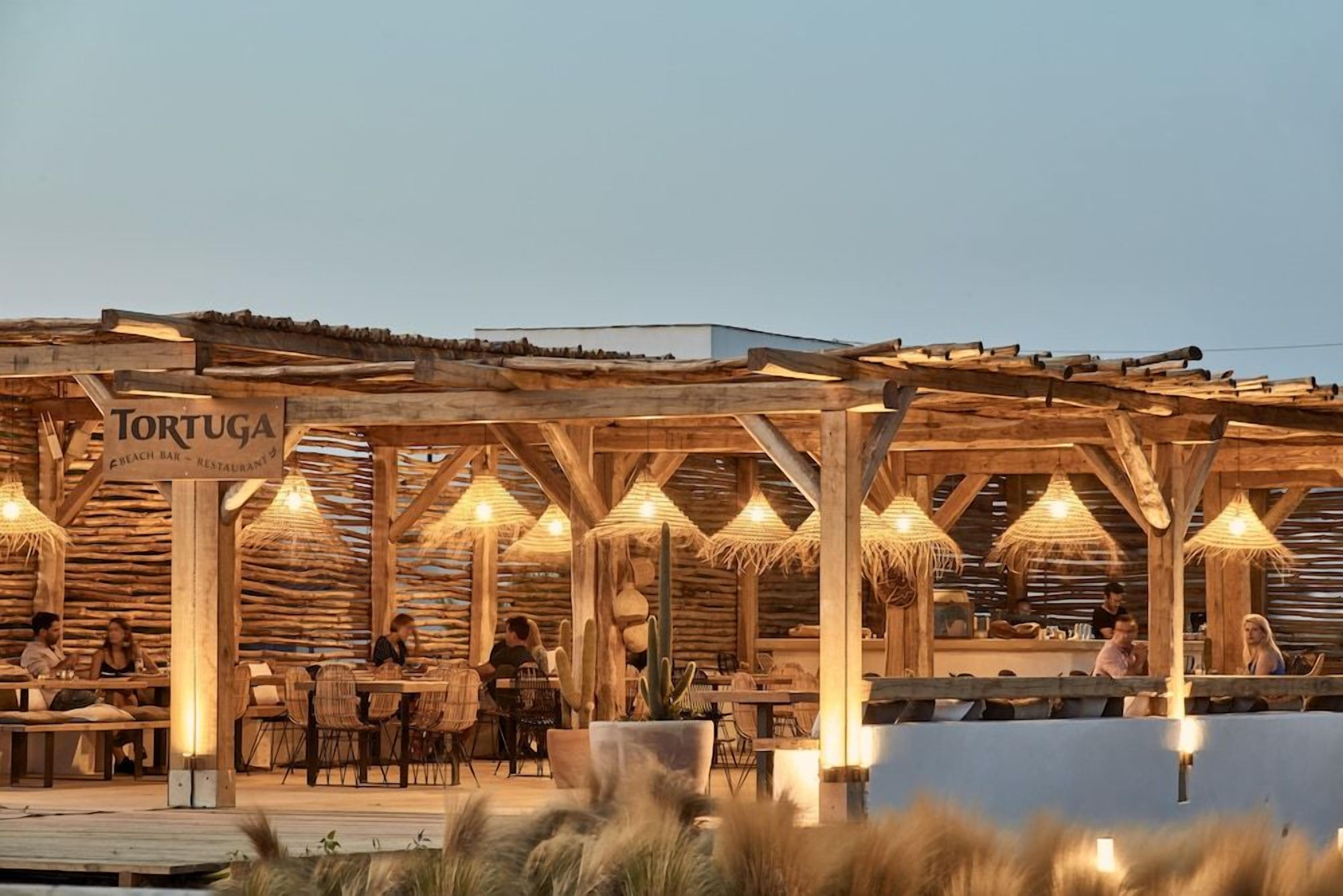 An outdoor dining area with wooden structures and tables, a sign reading 'TORTUGA BEACH BAR - RESTAURANT,' and a view of the sea in the background.