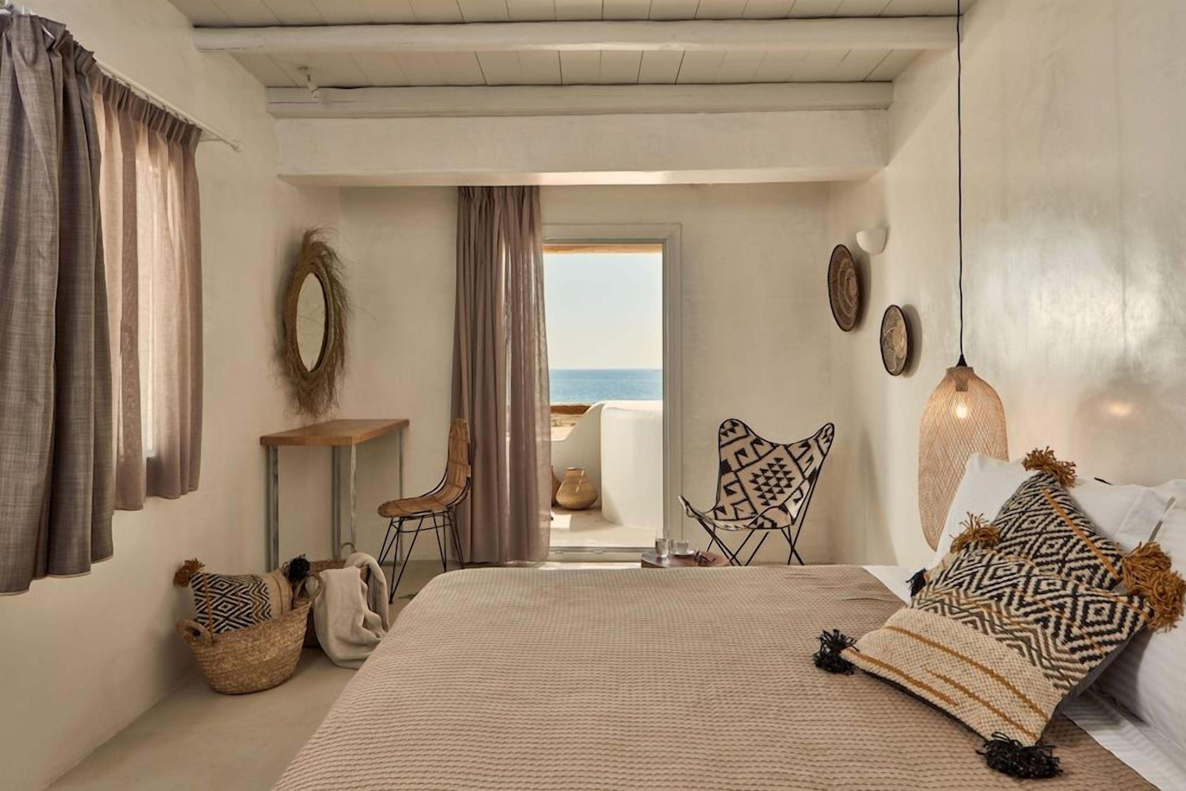 A bedroom with a bed, a chair, a small table, and a view of the sea through a doorway.