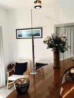 Una sala de estar moderna con una mesa de comedor de madera, una lámpara de pie, un cuadro en la pared, un jarrón con flores y una silla con cojín.