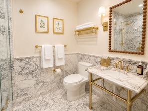 A bathroom with a marble floor, a toilet, a sink with gold fixtures, and two framed pictures on the wall. - Soju's Room at Keystone Hotel - 205 (Savannah)