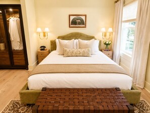 A neatly made bed with a headboard, bedside tables with lamps, a wooden bench, and a framed picture on the wall. - Soju's Room at Keystone Hotel - 205 (Savannah)