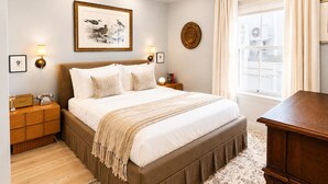 Een slaapkamer met een groot bed, nachtkastjes, een lamp, een lijst met een foto aan de muur, een raam met gordijnen en een houten dressoir.
