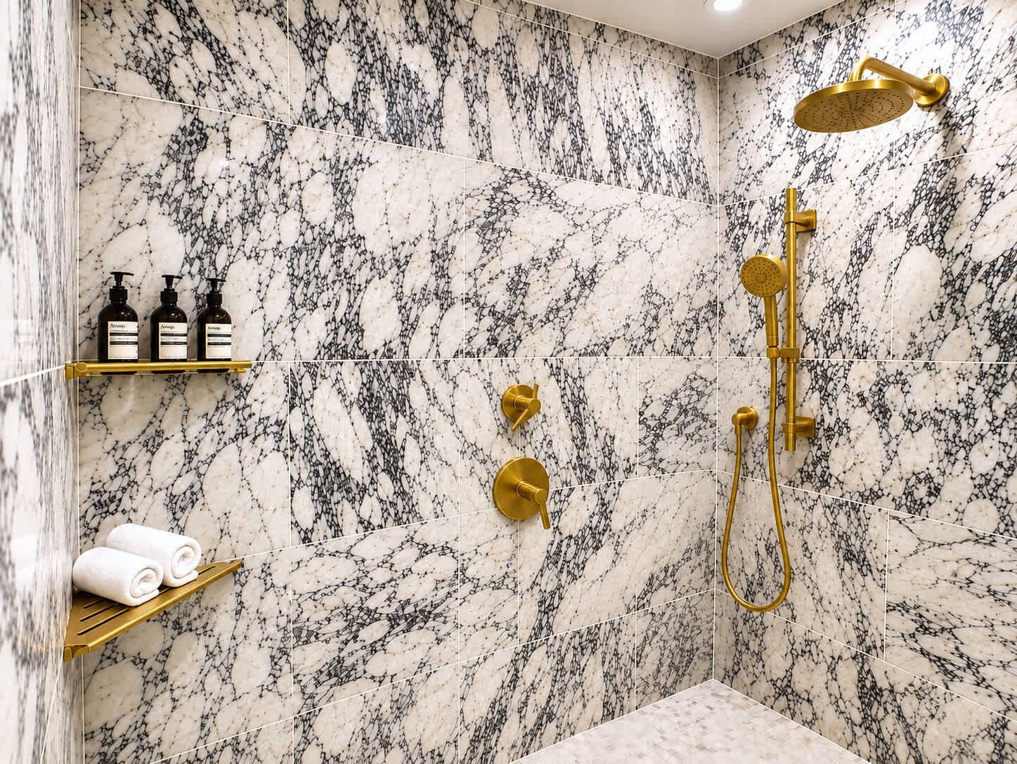 A bathroom with a marble shower wall, gold fixtures, and a shelf with toiletries.