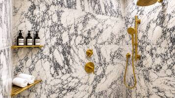 A bathroom with a marble shower wall, gold fixtures, and a shelf with toiletries.