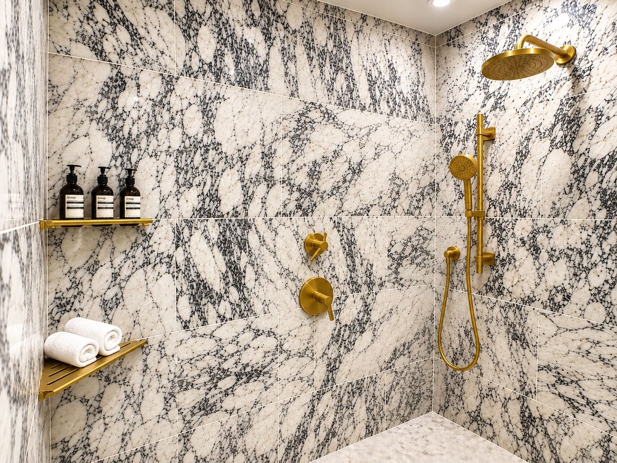 A bathroom with marble walls, a gold showerhead, and toiletries on a shelf.