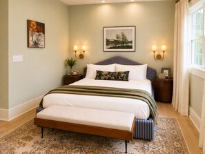 A bedroom with a large bed, bedside lamps, a nightstand, a window with curtains, and a painting on the wall.