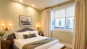 Ein ordentlich eingerichtetes Schlafzimmer mit einem großen Bett, Nachttischlampen, einem Fenster mit Vorhängen und einem gerahmten Bild an der Wand.